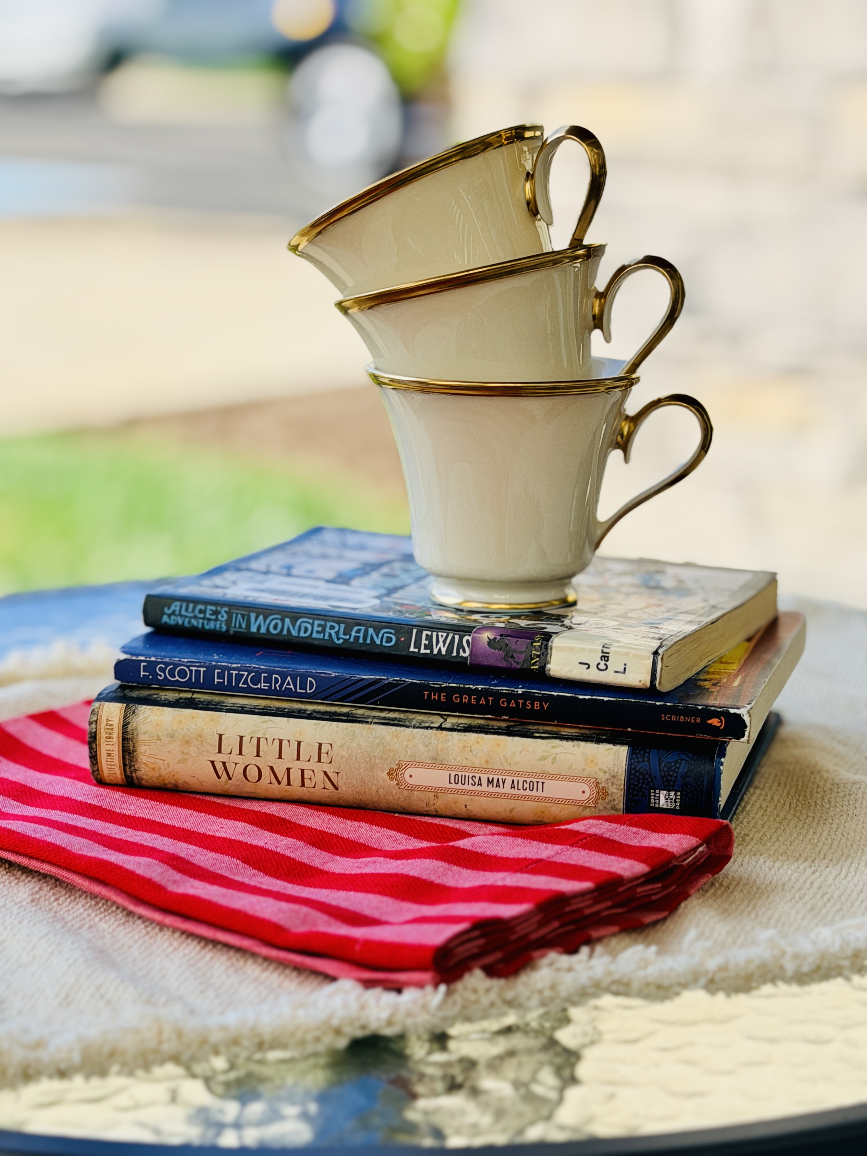 Gold-rimmed vintage teacups stacked atop a tower of classic novels including Little Women and The Great Gatsby
